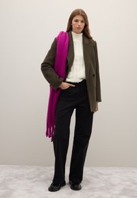 Abrigo de lana color verde oliva, suéter blanco de punto, pantalones negros de pierna ancha, bufanda fucsia sobre el hombro, zapatos negros gruesos.