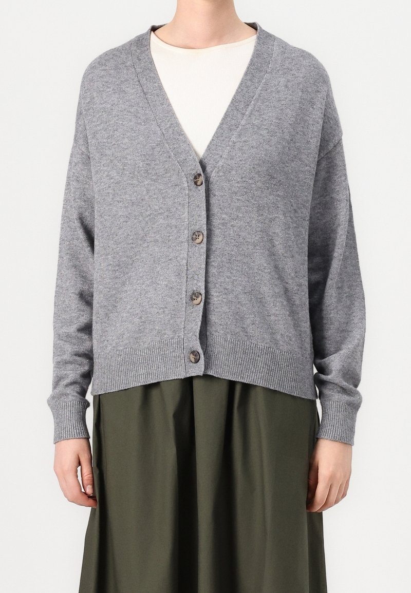 Personne vêtue d’un gilet gris boutonné par-dessus un haut beige clair et une jupe vert olive, debout devant un fond blanc uni.