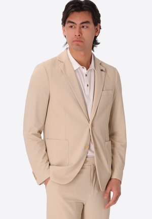 Uomo con giacca e pantaloni beige e polo bianca, in piedi con una mano in tasca, che guarda leggermente di lato.