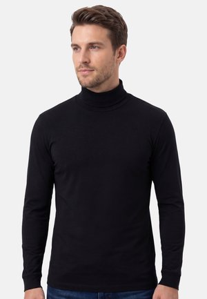 Mann mit hellbraunen Haaren, der einen schwarzen Rollkragenpullover mit langen Ärmeln und blaue Jeans trägt, leicht nach links auf weißem Hintergrund blickend.