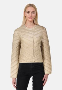 Ljust beige kviltad jacka med rund hals, horisontellt chevronmönster, tryckknappsstängning och långa ärmar. Bärs över svarta byxor.