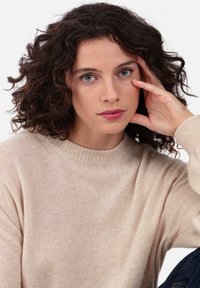 Pull beige en maille avec un col rond et des poignets côtelés. Le modèle a des cheveux bouclés et une expression neutre, posant sa main sur son visage.