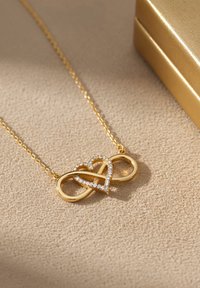 Guldhalskæde med et uendelighedssymbol flettet sammen med et hjerte, pyntet med klare krystaller, hvilende på en beige overflade.