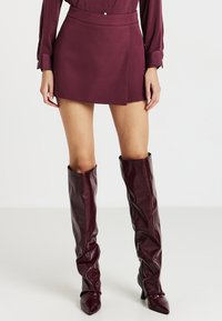 Donna che indossa una mini gonna bordeaux, una blusa a maniche lunghe abbinata e stivali in pelle con punta a punta fino al ginocchio.