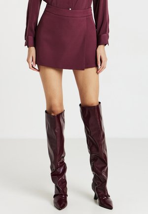 Donna che indossa una mini gonna bordeaux, una blusa a maniche lunghe abbinata e stivali in pelle con punta a punta fino al ginocchio.