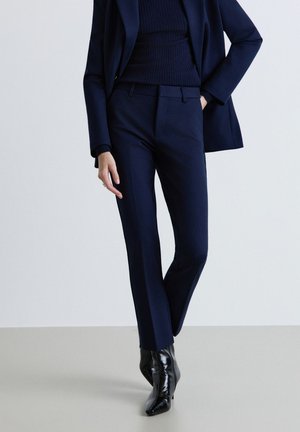 Persona che indossa pantaloni aderenti da completo blu navy, blazer abbinato, maglione a coste e stivaletti neri con punta appuntita su sfondo semplice.