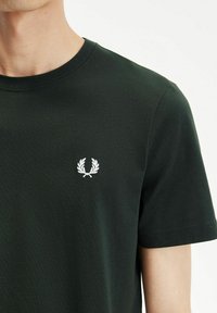 Camiseta de algodón verde oscuro con cuello redondo y mangas cortas. Presenta un logotipo bordado en blanco de laurel en el pecho. Textura suave.