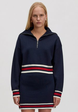 Marine blauwe ritsjurk met lange mouwen, met een gestreepte tailleband in rood, wit en blauw. Zachte, textuurstof.