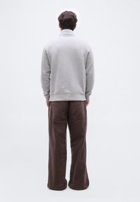 Άνδρας που στέκεται πλάτη, φοράει ανοιχτό γκρι φούτερ και χαλαρό καφέ παντελόνι μπροστά σε απλό λευκό φόντο.