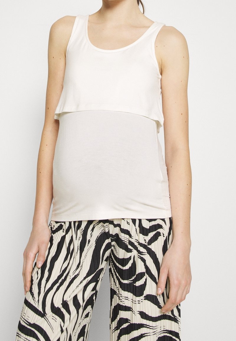 Femme portant un haut de maternité blanc sans manches et un pantalon plissé à taille haute imprimé zèbre noir et blanc, debout devant un fond uni.