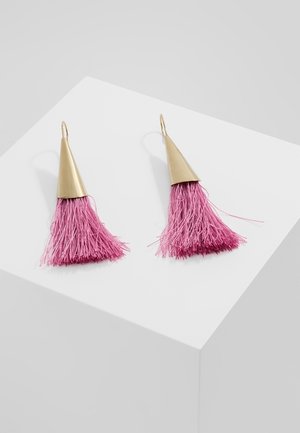 Kolczyki w kształcie stożków w złotym kolorze z długimi różowymi frędzlami z nici, prezentowane na białej ukośnej powierzchni.