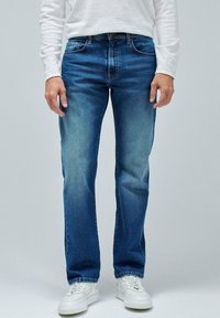 Vaqueros de denim azul con corte recto, lavado medio, cinco bolsillos y costuras amarillas, combinados con zapatillas blancas y una camisa de manga larga.