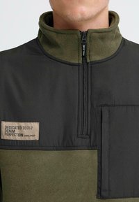 Fliisist pullover, millel on luku ees, tume roheline ülemine osa ja oliiviroheline fliisist alaosa; sisaldab plaastrilogot ja küljetaskut.