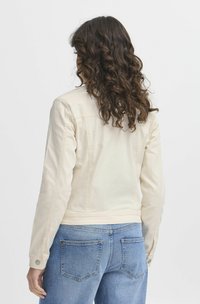 Veste en denim beige clair à la coupe ajustée, avec de longues manches et des boutons-pression. Associée à un jean bleu, mettant en valeur un style décontracté.