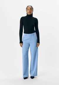 Schwarzer Rollkragenpullover kombiniert mit hellblauen weit geschnittenen Hosen. Das Oberteil hat eine taillierte Passform; die Hosen verfügen über Vorderfalten und eine glatte Textur.