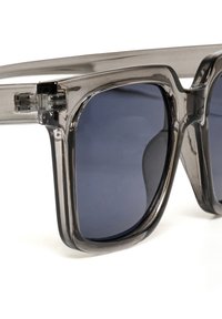 Lunettes de soleil grises transparentes avec des verres teintés foncés. Monture carrée épaisse avec une finition lisse et des charnières visibles.
