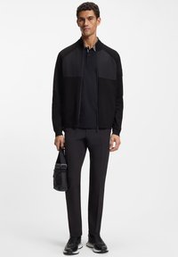 Μαύρο τζάκετ με φερμουάρ και υφασμένους ώμους, συνδυασμένο με μαύρο πόλο, ραμμένα παντελόνια και μαύρα αθλητικά παπούτσια, κρατώντας μια μικρή τσάντα.