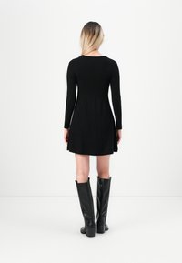 Femme aux cheveux blonds portant une robe noire à manches longues et des bottes noires montant jusqu'aux genoux, debout face à un fond blanc uni.