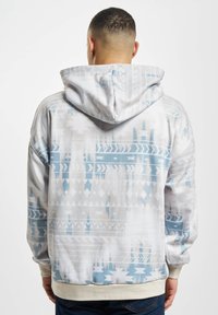 Sudadera con capucha en blanco con patrones geométricos en azul claro y gris. Tela suave, puños y dobladillo acanalados. Corte casual con una capucha amplia.