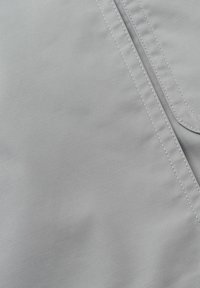 Tissu gris clair avec une couture double visible et une partie d'une poche à bords arrondis.