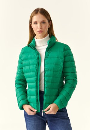 Giacca puffy verde con motivo trapuntato, colletto alto e chiusura con zip, indossata sopra un dolcevita bianco e abbinata a jeans blu.