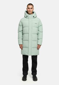 Giacca lunga imbottita verde chiaro con cappuccio, dotata di zip frontale, dettagli neri e sezioni a righe orizzontali. Indossata su pantaloni neri e stivali.