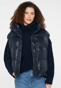Navy blue puffer vest with a shiny finish, zip closure, and snap buttons. Features a high collar and is worn over a dark sweater.