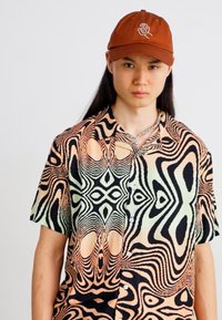 Chemise à manches courtes avec un motif noir et beige tourbillonnant, rehaussée de nuances vertes ; associée à une casquette marron ornée d'un logo.
