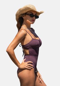 Maillot de bain une pièce texturé violet avec des bretelles fines et des côtés découpés. Assorti à un chapeau en paille et des lunettes de soleil noires, avec un collier superposé.