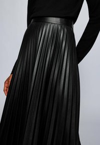 Gonna nera a pieghe realizzata in pelle sintetica. Presenta una vita ampia e liscia con pieghe verticali profonde lungo tutta la lunghezza della gonna.