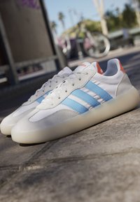 Hvide Adidas sneakers med lyseblå striber og orange hæftemærker placeret på en stensurface med en sløret udendørs baggrund.