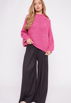 Flauschiger rosa Pullover mit hohem Kragen, weiten Ärmeln und asymmetrischem Saum, kombiniert mit schwarzen Hosen mit weitem Bein. Weiche Textur, entspannter Stil.