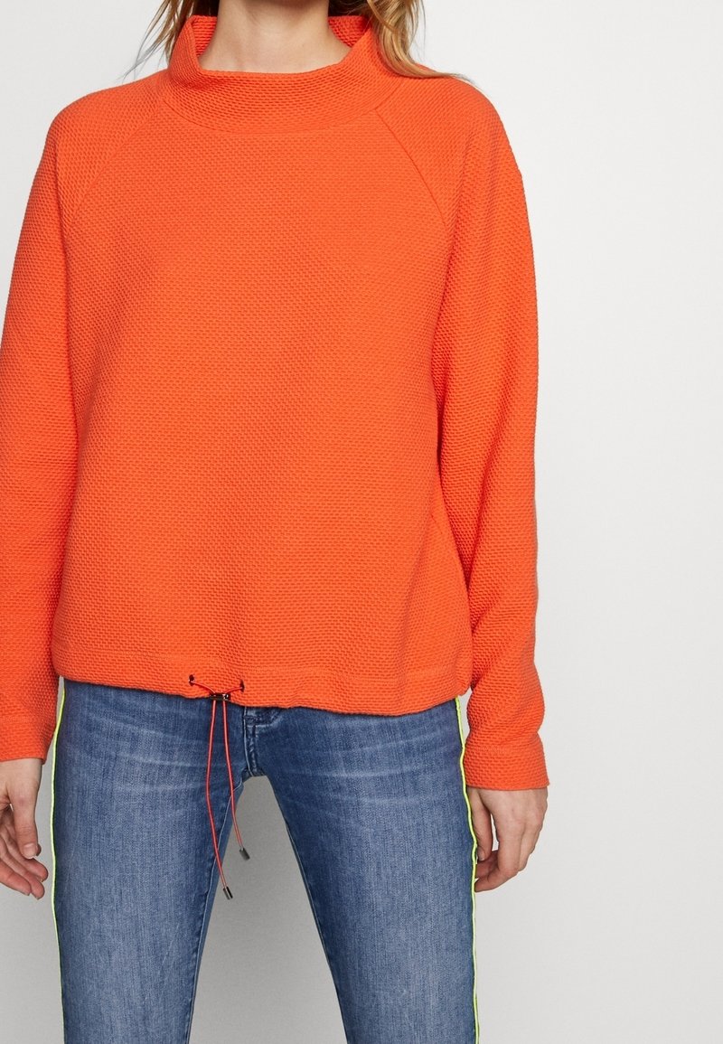 Orange, strukturierter Pullover mit hohem Kragen und Kordelzugsaum. Kombiniert mit blauen Jeans mit gelben Seitestreifen.