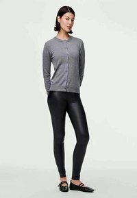Cárdigan gris con botones, combinado con leggings negros brillantes. El modelo lleva zapatos planos negros. Diseño simple y ajustado, con textura suave en toda la prenda.
