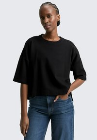 T-shirt noir cropped avec des manches courtes, un col rond et des fentes sur les côtés. Porté avec un jean bleu, doté d'une taille cintrée et d'une jambe droite.