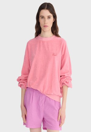 Kobieta z długimi brązowymi włosami nosi luźną różową bluzę z podwiniętymi rękawami i jasnofioletowe szorty, stoi na tle jednolitego jasnego tła.