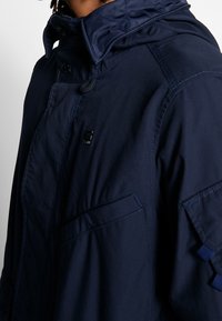 Man som bär en marinblå jacka med hög krage, knäppning fram och ficka på armen, visad från bröstet till nacken.