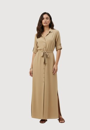 Femme portant une robe maxi beige boutonnée devant avec manches retroussées, ceinture à la taille, fentes latérales, et sandales plates marron sur un fond uni.
