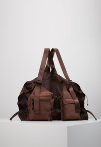 Brun nylon gympapåse med justerbara remmar, två sidofickor och dragkedjedetaljer. Mjuk textur och minimalistisk design.