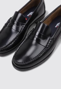 Los mocasines de cuero negro presentan un acabado liso, detalles cosidos y un diseño clásico con un panel delantero elevado. La suela tiene textura.