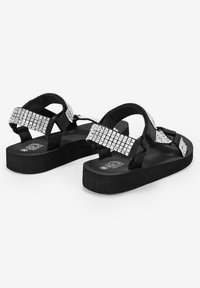 Sandalias negras con correas texturizadas que presentan un diseño de lentejuelas plateadas. Suela de goma plana, accesorio de hebilla ajustable y estilo destapado.