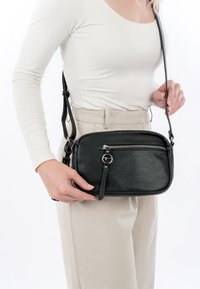 Frau trägt beige Hose und weißes Langarmshirt, hält eine schwarze Leder-Umhängetasche mit vorderer Reißverschlusstasche.