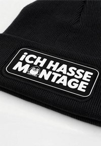 Black knitted beanie with a folded cuff, featuring a rectangular patch that reads "iCH HASSE MONTAGE" in white, with a cartoon penguin graphic.