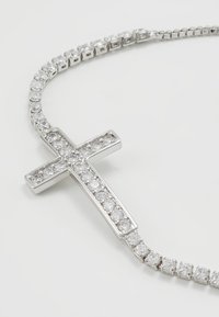 Sidabrinės spalvos kaklo grandinėlė su kryžiaus pakabuku, puoštu skaidriais, apvaliais cirkoniais, uždėta ant grandinėlės su pakaitiniais kvadratiniais jungtimis.
