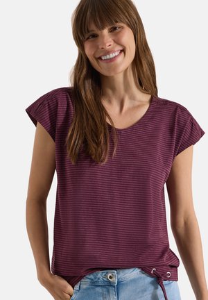 Femme souriante aux longs cheveux bruns porte un top rayé bordeaux à manches courtes avec un lien sur le côté, associé à un jean bleu clair.