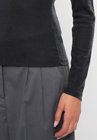 Μαύρη μπλούζα με μακριά μανίκια από υφασμάτινη υφή με ασημένιες λεπτομέρειες, συνδυασμένη μ�ε πλισέ γκρίζες παντελόνες. Κοντινό στο χέρι και το χέρι.