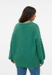 Pull vert surdimensionné avec une finition en tricot texturé, des poignets côtelés et un dos présentant des accents en tricot torsadé. Associé à un jean en denim bleu.