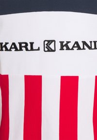 Červenobíle pruhovaná látka s logem "KARL KANI" v černé barvě. Horní část je jednobarevná tmavě modrá s strukturovanou texturou.