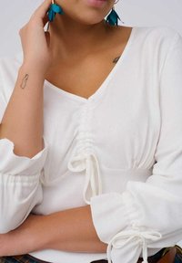 Witte blouse met een V-hals, geplooide voorkant en lange, puffed mouwen. De stof lijkt soepel; heeft trekkoordaccenten bij de polsen.