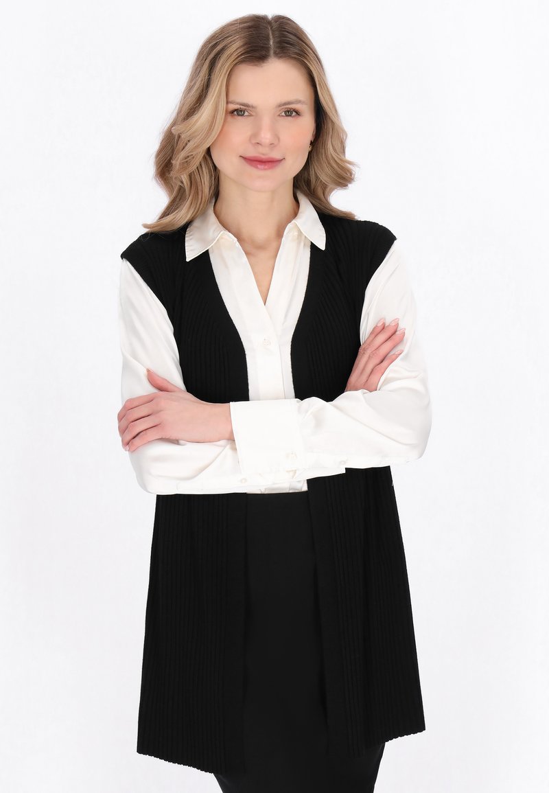 Blonde vrouw met gevouwen armen, gekleed in een witte blouse en een zwart mouwloos vest, staand tegen een witte achtergrond.